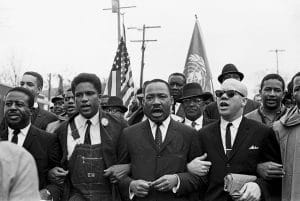 A Black and white photograph from the Selma to Montgomery marches. In the center is Martin Luther King Jr, arms linked and walking with protesters.